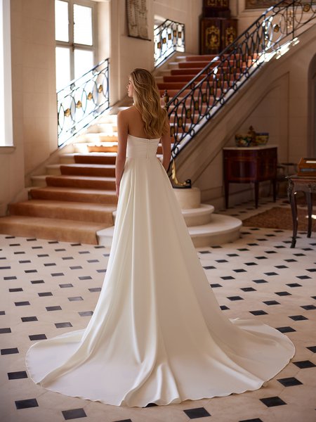 Back view of a bride in a luxurious ivory crepe wedding dress, featuring an open back and flowing chapel train, before a staircase Moonlight Couture H1601