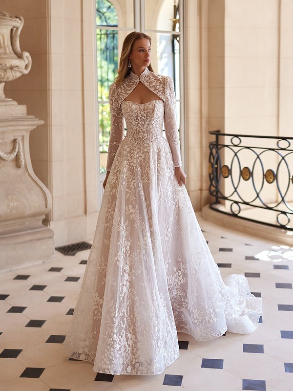 A stunning bride in Moonlight Couture H1605 a luxurious lace ball gown poses in an elegant hallway, featuring a sweetheart bodice and pearl details