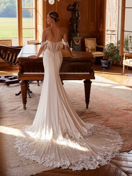 A bride in a luxurious crepe wedding dress with lace detailed train stands poised by a piano, radiating elegance and romance on her special day Moonlight Couture H1606