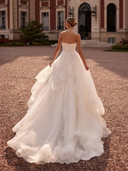 A bride in a stunning open-back wedding dress adorned with lace and pearls walks elegantly in front of an impressive structure Moonlight Couture H1607