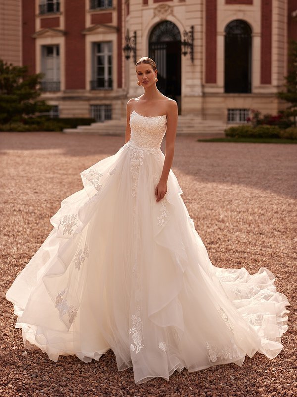 A bride in a scoop neck A-line cascade skirt wedding dress adorned with lace and pearls stands gracefully before an elegant mansion Moonlight Couture H1607