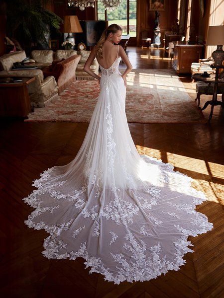 A bride is seen from behind in an opulent room, showcasing the back of her elegant ivory wedding gown. The dress features a low, deep scoop back, thin straps, and intricate floral lace details cascading down the form-fitting bodice and flowing into the sheer, chapel-length train. The lace-trimmed train spreads gracefully on the wooden floor, creating a dramatic effect.