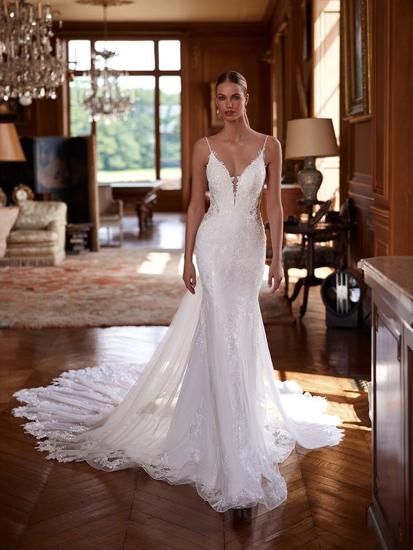 A bride stands in a luxurious, sunlit room wearing a stunning, form-fitting ivory wedding gown. The dress features a deep V-neckline with delicate lace straps, intricate floral lace detailing throughout, and a long, flowing train with sheer fabric and lace embellishments.