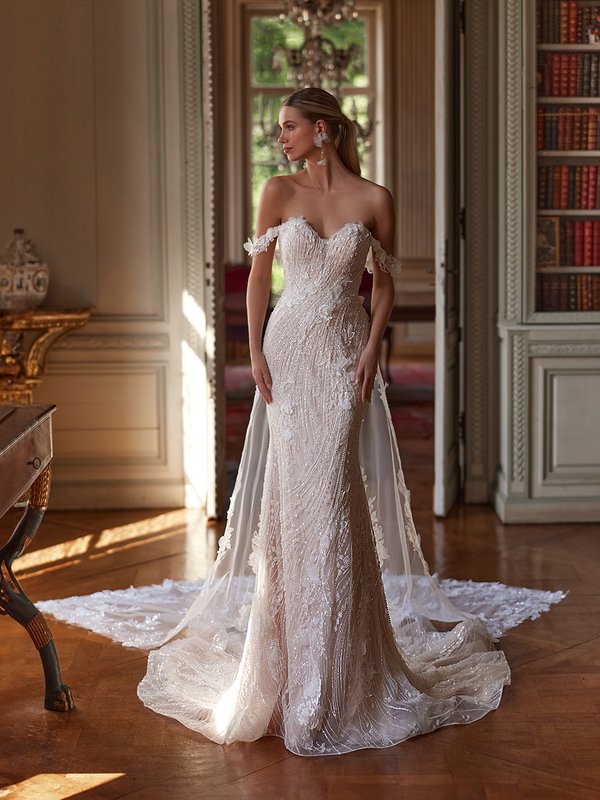 A bride in an ornate room wearing an off-the-shoulder, beaded wedding gown with a long train, standing with her hands at her sides.