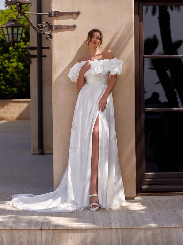 A bride in an elegant off-the-shoulder ivory jacquard bridal gown is standing outdoors against a beige wall, with soft shadows cast across her. The dress features a voluminous off the shoulder ruffled jacket and a thigh-high slit, revealing her left leg. The setting is sunlit and lush green foliage in the background, giving a romantic, Mediterranean feel to the scene.