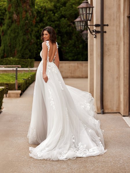 A bride poses outdoors, showcasing the back view of her elegant wedding gown. The dress features a deep V-back with delicate floral lace appliqué along the edges of the straps and down the sides, adding a romantic, ethereal touch. The voluminous A-line skirt flows gracefully behind her, creating a long train adorned with scattered floral lace appliqués near the hemline.