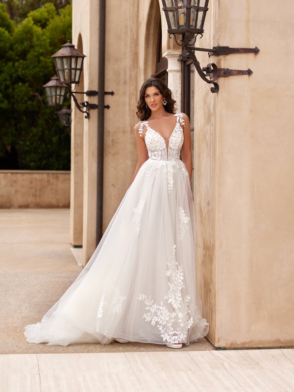 Bride in wedding dress that has a sheer bodice with lace patterns overlaid, extending to the wide straps adorned with floral embellishments. The flowing A-line skirt features layers of soft tulle with lace appliqués concentrated near the hem, adding a romantic touch.