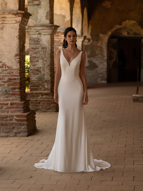 bride stands in an elegant, minimalist white wedding dress with a fitted bodice featuring a deep V-neckline and wide shoulder straps. The gown has a smooth, tailored silhouette that flows into a subtle train.